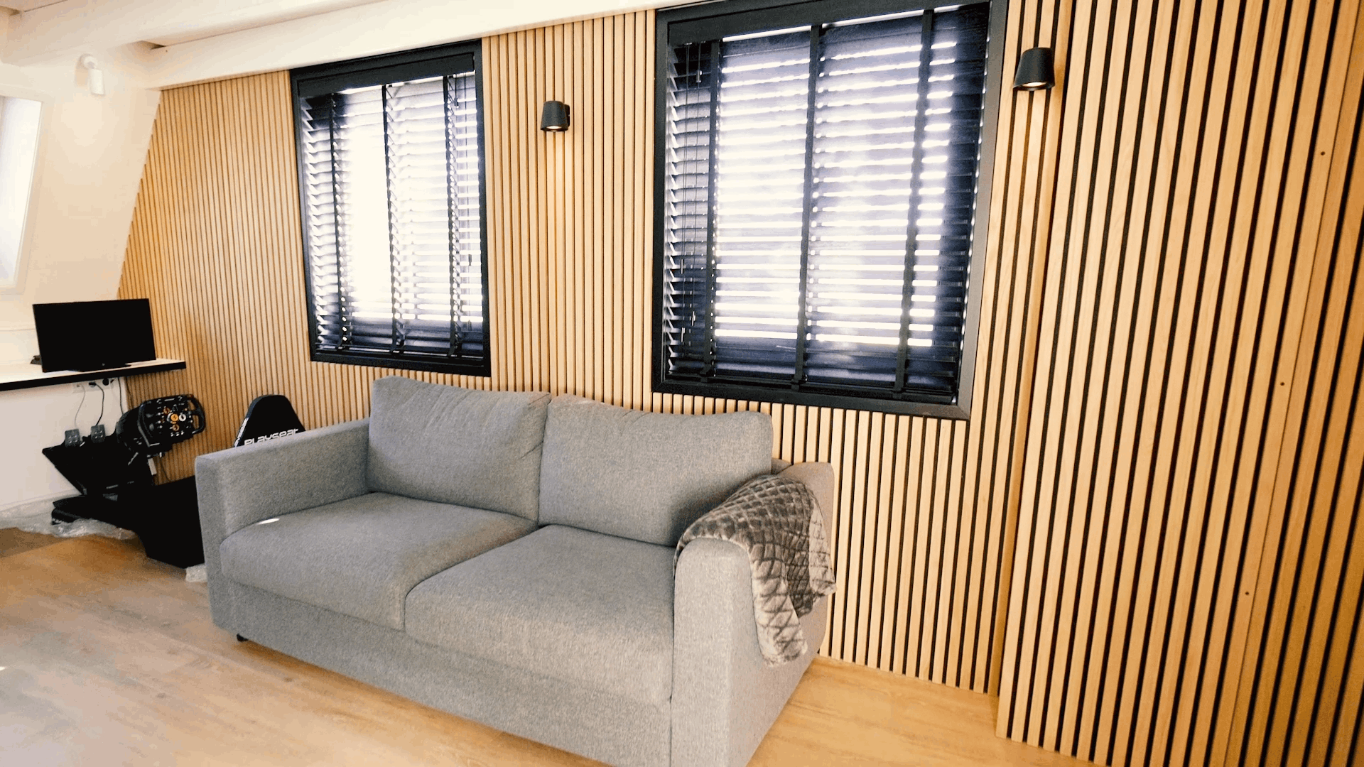 Graues Sofa mit gemusterter Decke vor einer nordischen Akupanel-Akustik-Holzlattenwand, flankiert von zwei Fenstern mit schwarzen Jalousien und einer kompakten Schreibtischeinrichtung.